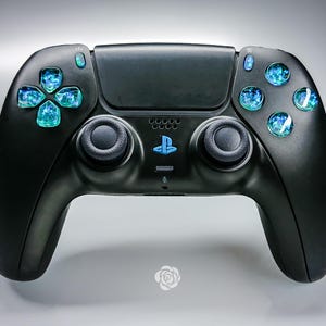 Pode incluir: Controlador de jogos preto com botões e direcional azuis brilhantes. O controlador tem dois manípulos analógicos pretos e um símbolo PlayStation azul. Os botões têm uma aparência texturizada semelhante a uma gema.