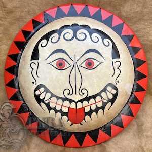 May include: A round, painted shield featuring a stylized face with red eyes, a long nose, and a protruding red tongue. The face is surrounded by a black and red triangular border. The shield has a cream-colored background.