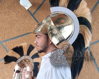 Puede incluir: Un casco espartano de color dorado con una pluma negra y beige. El casco lo sostiene una persona que lleva una camisa blanca. También es visible un segundo casco más pequeño. El fondo presenta una pared texturizada.