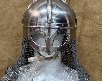 May include: A polished silver Viking helmet with a face guard and chainmail. The helmet is displayed on a white mannequin head against a brown background. The helmet has a central ridge and is riveted.