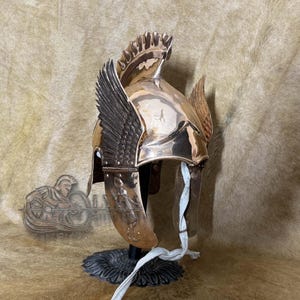 May include: A bronze-coloured helmet with wing accents and a decorative crest. The helmet features a detailed design, including a wing on each side and a crest at the top. It is displayed on a black stand with a white strap.