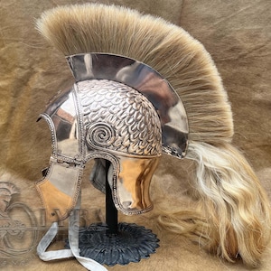 May include: An antique silver-coloured helmet with a large, light brown plume. The helmet has a detailed, scale-like pattern on the crown and a decorative swirl design. It is displayed on a black stand.