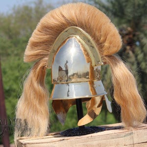 May include: A silver and gold-toned helmet with a long, flowing, light brown mane. The helmet is displayed on a wooden surface, showcasing its intricate design and historical aesthetic. The helmet's polished surface reflects the surrounding environment.