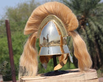 Puede incluir: Un casco plateado y dorado con una larga melena rubia y fluida. El casco se exhibe sobre una superficie de madera, mostrando su intrincado diseño y estética histórica. La superficie pulida del casco refleja el entorno.