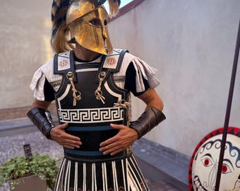 Puede incluir: Un casco espartano dorado con una cresta de plumas, usado con una coraza y falda en blanco y negro. La armadura presenta diseños intrincados y se combina con brazales y botas de cuero marrón. También se ve un escudo redondo con un diseño de rostro y una lanza.