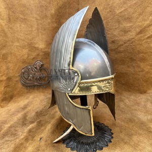 Peut inclure: Casque argenté avec des extensions en forme d'ailes et des bordures dorées. Le casque, orné d'une crête décorative, est présenté sur un socle noir. Le design évoque un thème historique ou fantastique.