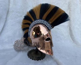 Puede incluir: Un casco espartano de color cobre pulido con una pluma negra y marrón. El casco presenta una banda decorativa con un patrón de clave griega y se exhibe sobre un soporte negro. Es una réplica histórica.