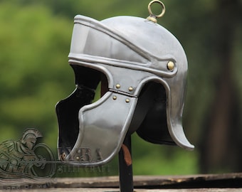 May include: A silver-colored Roman helmet with a rounded top, visor, and cheek guards. The helmet has a brass ring on top and is riveted with small brass studs. The background is a blurred green.