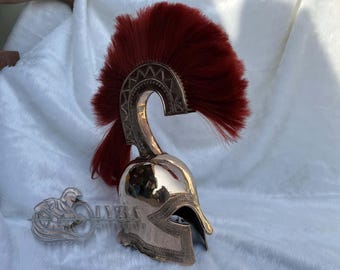 Puede incluir: Un casco espartano de color bronce con una pluma roja. El casco tiene detalles intrincados a lo largo de la frente y una cresta curva. Un emblema decorativo de metal con una figura está al lado del casco.