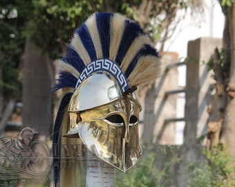Puede incluir: Un casco espartano de latón pulido con una pluma azul y beige. El casco presenta ranuras para los ojos y una banda decorativa alrededor de la frente. La pluma tiene rayas azules y beige alternas.