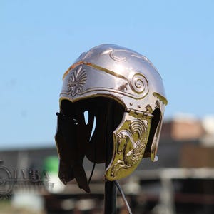 Puede incluir: Un casco plateado y ornamentado con detalles dorados. El casco presenta grabados detallados, incluyendo un diseño floral y un caballo alado. El casco es probablemente un accesorio histórico o teatral.