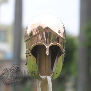 May include: A polished copper-coloured helmet with a rounded top and a decorative front. The helmet has a reflective surface and is mounted on a wooden post. The design includes cheek guards and a detailed brow. The background is blurred, suggesting an outdoor setting.