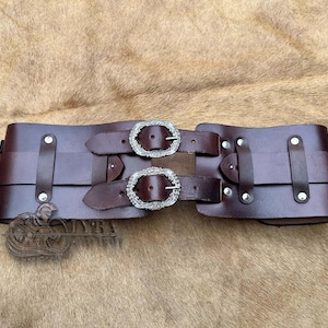 May include: A wide, dark brown leather belt with two ornate silver buckles. The belt has multiple straps and silver rivets. The design suggests a medieval or fantasy theme, suitable for costumes or historical reenactments.