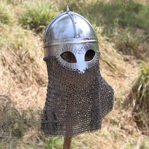 May include: A silver Viking helmet with a chainmail face guard. The helmet has eye openings and is riveted. The chainmail extends down to cover the neck and shoulders. The helmet is displayed outdoors against a backdrop of grass and foliage.