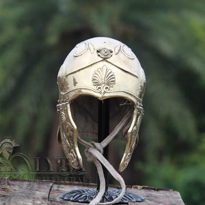 May include: A gold-coloured, ornate helmet with detailed carvings. The helmet features a decorative crest and cheek guards, and is displayed on a stand with a neutral background. The helmet's design suggests a historical or fantasy theme.