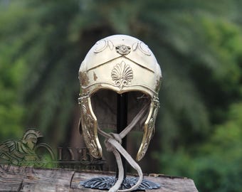 Puede incluir: Un casco dorado y ornamentado con tallas detalladas. El casco presenta una cresta decorativa y protectores para las mejillas, y se exhibe sobre un soporte con un fondo neutro. El diseño del casco sugiere un tema histórico o de fantasía.