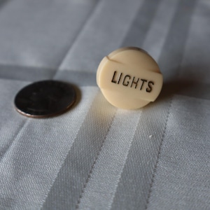 May include: A round, off-white plastic knob featuring a raised horizontal bar with the word LIGHTS embossed in black lettering. The knob is positioned next to a standard United States quarter coin for scale, showing its small, compact size.
