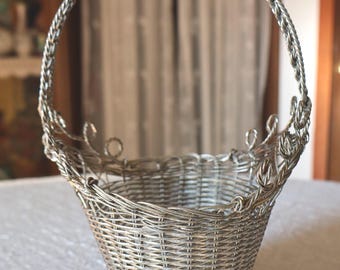 May include: A silver-colored wire basket featuring a woven body, a flared circular base with a lattice pattern, and a tall, twisted wire handle. The upper rim is decorated with small, looped wire accents that extend upward from the basket edge.