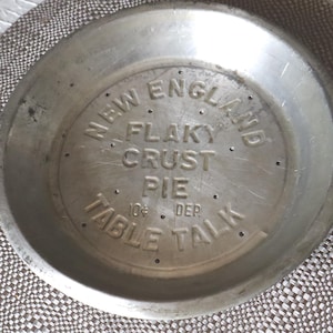 May include: A vintage, round, aluminum pie pan with the words "NEW ENGLAND FLAKY CRUST PIE TABLE TALK" embossed on the inside. The pan has small holes and is sitting on a patterned placemat.