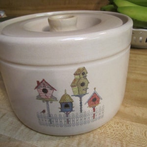 May include: A cream-colored ceramic container with a lid. The container features a colorful illustration of birdhouses on a white picket fence. The lid has a small, rounded handle. The container is likely for kitchen storage.
