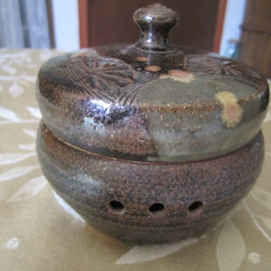 May include: A brown ceramic jar with a lid, featuring a speckled glaze and a decorative design. The lid has a knob and a floral pattern.