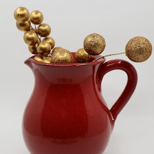 May include: A red ceramic pitcher with a curved handle, filled with gold-colored decorative spheres. Some spheres are clustered, while others are on thin gold stems, creating a festive arrangement. The pitcher has a glossy finish.
