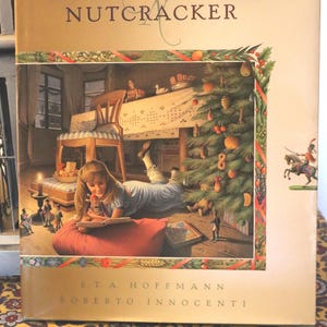 May include: A hardcover book titled "Nutcracker" with a gold cover. The cover features an illustration of a young girl reading near a Christmas tree. The author's name, E.T.A. Hoffmann, and the illustrator's name, Roberto Innocenti, are printed on the bottom.