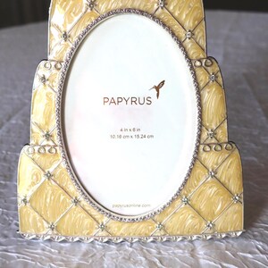 May include: A decorative yellow photo frame with an oval opening. The frame has a quilted pattern and is embellished with rhinestones. The text "PAPYRUS" and "4 in x 6 in" are visible. The frame measures 10.16 cm x 15.24 cm.
