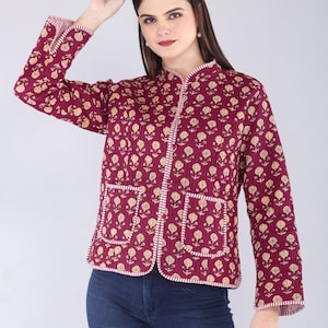 May include: Burgundy quilted jacket with a floral pattern in gold. The jacket has a stand-up collar, two front pockets, and white trim. The sleeves are long, and the jacket is buttoned up the front.
