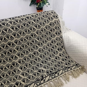 May include: A black and beige patterned throw blanket with fringe. The blanket features a repeating fan-like design. The throw is draped over a white couch. A vase of flowers sits in the background.