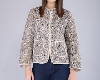 May include: A beige quilted jacket featuring a black floral print, a round neckline, long sleeves, and two front patch pockets with white stitched trim. The jacket is worn over dark blue denim jeans, creating a layered look.