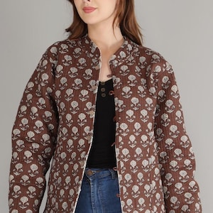 May include: A brown quilted jacket with a white floral pattern. The jacket has a mandarin collar and is buttoned up. The model is wearing a black top and blue jeans.