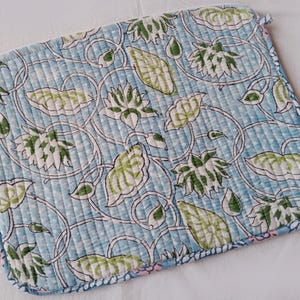 May include: A quilted, square potholder with a floral pattern. The design features white, green, and light blue flowers and leaves on a light blue background. The edges are finished with a blue binding and a small loop for hanging.