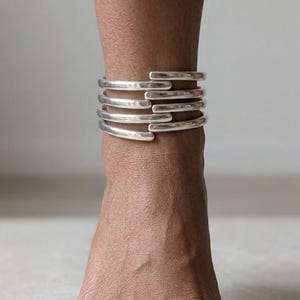 May include: A set of five silver-toned, open-ended anklets stacked on an ankle. The anklets have a smooth, polished finish and a simple, minimalist design. The skin tone is medium brown.