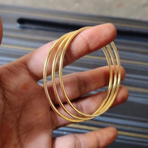 May include: A set of four thin, gold-colored bangles. The bangles are round and appear to be made of metal. The jewelry is held in a hand against a blurred background.