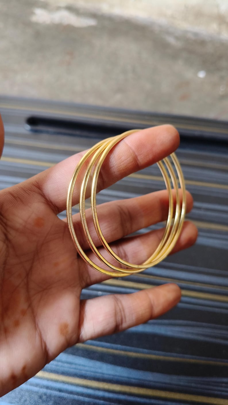 May include: A set of four thin, gold-colored bangles. The bangles are round and appear to be made of metal. The jewelry is held in a hand against a blurred background.