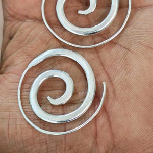 Puede incluir: Dos pendientes plateados en espiral. Cada pendiente presenta un diseño en espiral, con la forma espiral ensanchándose gradualmente desde el centro. Los pendientes se muestran en una mano, mostrando su tamaño y diseño. La joyería es simple y elegante.