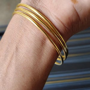 May include: A close-up of a wrist adorned with multiple thin, gold-colored bangles. The bangles are smooth and shiny, reflecting light. The skin tone is light brown, and the background is blurred.