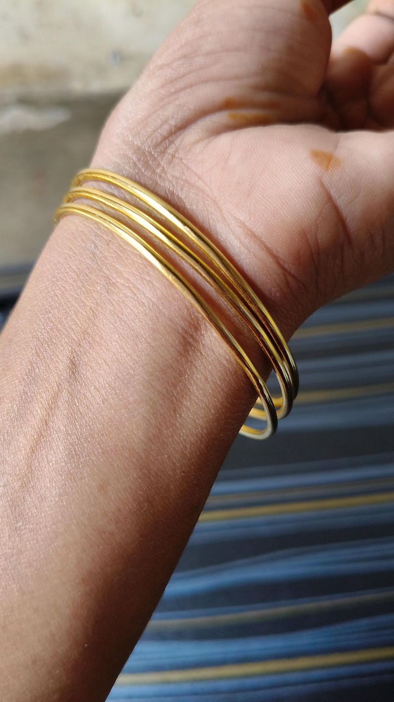May include: A close-up of a wrist adorned with multiple thin, gold-colored bangles. The bangles are smooth and shiny, reflecting light. The skin tone is light brown, and the background is blurred.