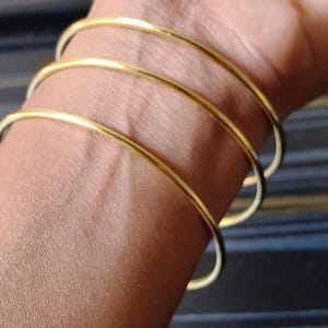 May include: Close-up of a wrist adorned with four thin, gold-colored bangles. The smooth, circular bracelets are stacked, creating a layered effect. The skin tone is a warm, medium shade, contrasting with the bright metal. The background is a dark, striped pattern.