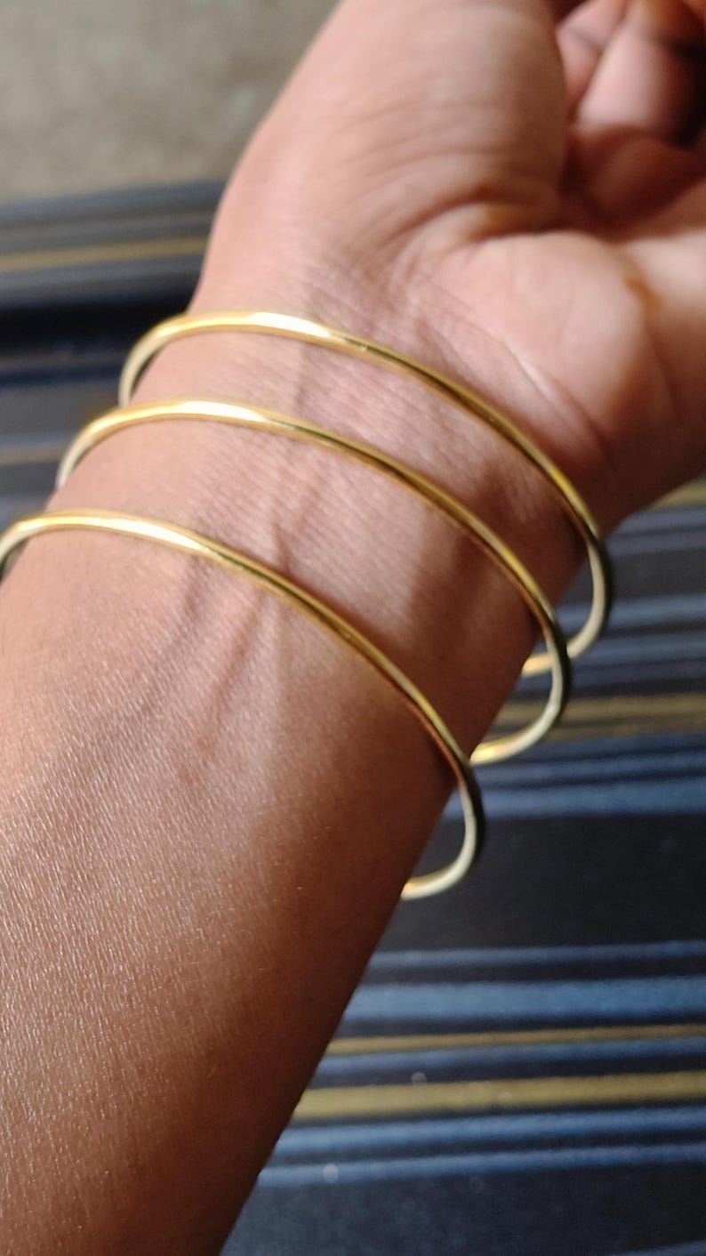 May include: Close-up of a wrist adorned with four thin, gold-colored bangles. The smooth, circular bracelets are stacked, creating a layered effect. The skin tone is a warm, medium shade, contrasting with the bright metal. The background is a dark, striped pattern.