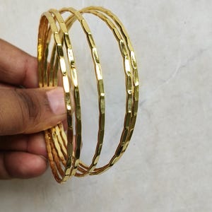 May include: A set of gold-colored bangles with a textured, hammered design. The bangles are held in a hand, showcasing their shiny, metallic appearance. The jewelry is displayed against a light-colored background.