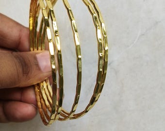 May include: A set of gold-colored bangles with a textured, hammered design. The bangles are held in a hand, showcasing their shiny, metallic appearance. The jewelry is displayed against a light-colored background.