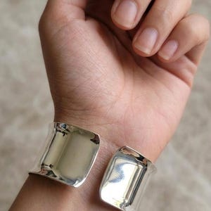 May include: A pair of wide, silver-colored cuff bracelets. The bracelets are open-ended and have a smooth, reflective surface. The image shows the bracelets on a wrist, with the light reflecting off the polished metal.