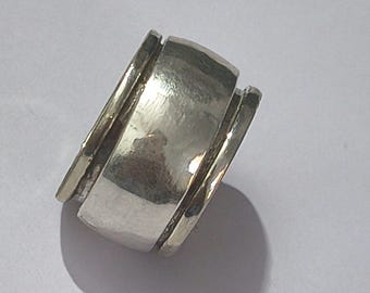 Könnte beinhalten: Ein silberner Ring mit breitem Band und strukturierter Oberfläche. Der Ring hat ein einzigartiges Design mit einem zentralen, leicht gebogenen Abschnitt, der von erhöhten, parallelen Kanten flankiert wird. Das Metall hat ein gehämmertes Aussehen und wirkt rustikal.