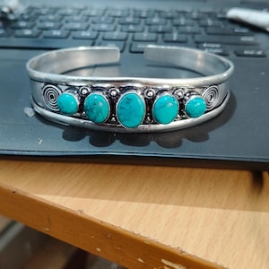 Puede incluir: Una pulsera de plata con cinco piedras turquesas. La pulsera tiene un diseño abierto y presenta patrones decorativos en espiral y remolino. Las piedras turquesas son de forma ovalada y están engastadas en biseles individuales. La pulsera está sobre una superficie negra.