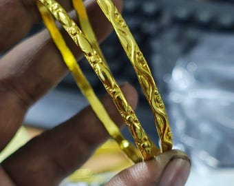 May include: Three gold bangles with intricate, embossed floral and swirl patterns. The bangles are held in a hand, showcasing their bright, reflective surface. The jewelry appears to be made of gold.