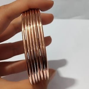 May include: A set of five thin, polished rose gold-toned metal bangles. Each bangle features a smooth, rounded, and reflective surface. The bracelets are stacked together, showcasing a uniform, metallic finish with a warm, pinkish-copper hue.