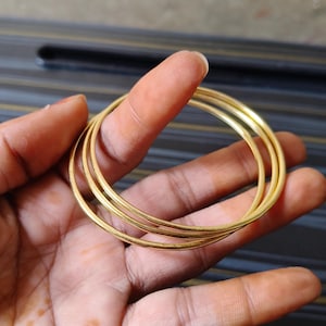 May include: A set of four gold-colored bangles. The bangles are thin and round, with a smooth, polished finish. The jewelry is held in a hand against a dark background.