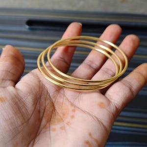 May include: A set of gold-colored bangles, stacked on top of each other, are held in a person's hand. The bangles are thin and have a smooth, reflective surface. The background is a blurred mix of colors.
